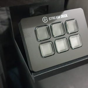 May include: A black Stream Deck with six illuminated square buttons. The device has the "STREAM DECK" logo at the top. It is mounted on a black stand, likely for desktop use. The buttons are clear and the device is rectangular.