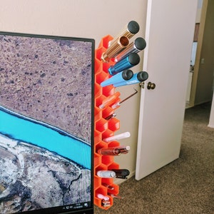 Op de afbeelding: Oranje honingraatvormige bureau-organizer met verschillende pennen, potloden en gereedschap. De organizer is bevestigd aan de zijkant van een computermonitor.