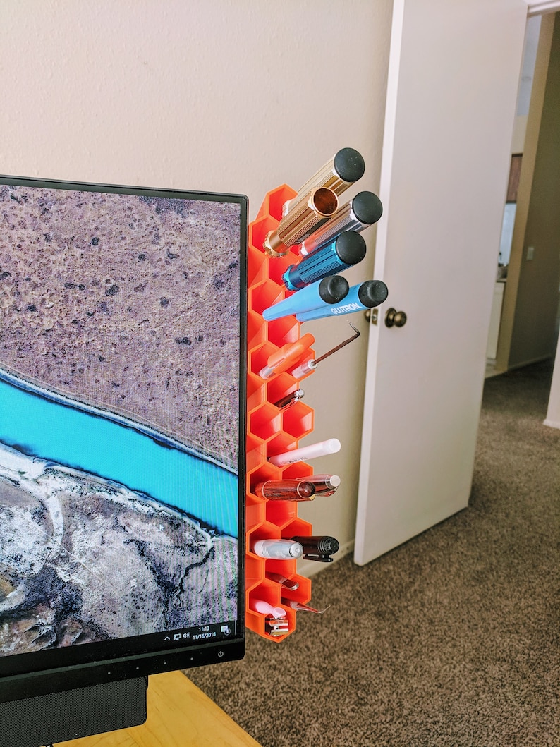 Op de afbeelding: Oranje honingraatvormige bureau-organizer met verschillende pennen, potloden en gereedschap. De organizer is bevestigd aan de zijkant van een computermonitor.