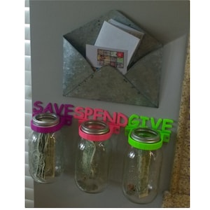 May include: Three glass jars with money inside, labeled "SAVE", "SPEND", and "GIVE" with pink and green plastic labels. The jars are mounted on a wall with a metal envelope attached above.