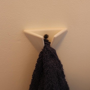 Puede incluir: Un gancho de pared triangular blanco con un tornillo oscuro que sostiene una toalla azul oscuro. El gancho está montado en una pared beige claro, ofreciendo un diseño simple y moderno para el baño o la cocina.
