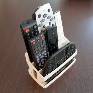 May include: A white plastic remote control organizer with six slots for remotes. The organizer is filled with five different remotes, including a black remote with the word "POWER" on it, a black and silver remote with the word "SKY" on it, and a black remote with a small screen and a keyboard.