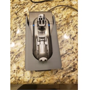 May include: A gray plastic charging dock for a cordless vacuum cleaner. The vacuum cleaner is plugged in and charging, indicated by the blue lights on the dock.