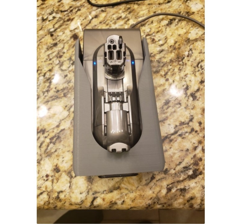 May include: A gray plastic charging dock for a cordless vacuum cleaner. The vacuum cleaner is plugged in and charging, indicated by the blue lights on the dock.