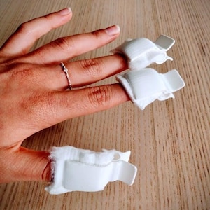 May include: A hand with a delicate ring on one finger, and three fingers with white finger splints. The splints are made of a soft, padded material and have a rigid, white plastic clasp.