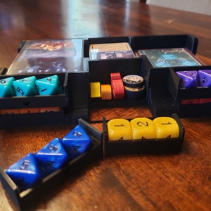 May include: A black plastic organizer with compartments for dice and cards. The organizer has a compartment for dice with a blue, green, and purple dice. The organizer also has a compartment for cards with a card that says "Haunted Woods".