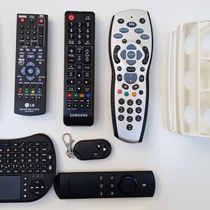 May include: A collection of eight different remote controls, including a black and silver LG Blu-ray Disc Player remote, a black Samsung TV remote, a silver Sky remote, a white Samsung TV remote, a black and gray wireless keyboard with a touchpad, a black Amazon Fire TV remote, and a small black remote with a keychain.