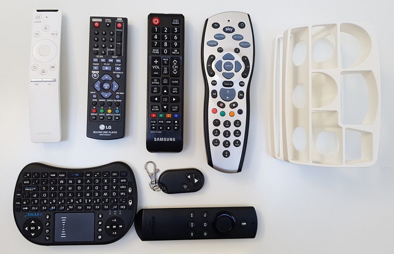 May include: A collection of eight different remote controls, including a black and silver LG Blu-ray Disc Player remote, a black Samsung TV remote, a silver Sky remote, a white Samsung TV remote, a black and gray wireless keyboard with a touchpad, a black Amazon Fire TV remote, and a small black remote with a keychain.