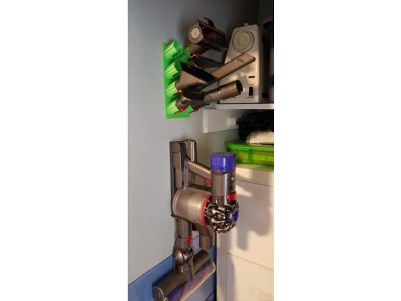 May include: A gray and black cordless vacuum cleaner with a blue and purple attachment is mounted on a wall-mounted holder. The vacuum cleaner is attached to a green wall-mounted storage rack with multiple attachments.