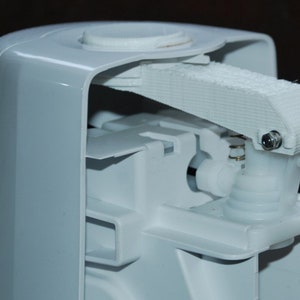 Pode incluir: Close-up de um dispensador de plástico branco com uma alavanca e um mecanismo de mola. O dispensador está aberto, revelando o funcionamento interno.