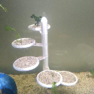 May include: A clear, tiered aquarium decoration with circular platforms filled with gravel and small plants. The structure is white and has a central column with several platforms extending outwards. The decoration is designed to be placed in an aquarium.
