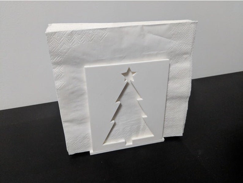 May include: White ceramic napkin holder shaped like a Christmas tree with a star on top. The holder is empty and ready to hold napkins.