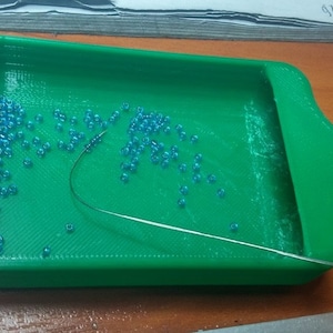 Könnte beinhalten: Ein grüner Plastik-Perlen-Organizer mit einem kleinen Ausguss. Der Organizer ist mit blauen Perlen gefüllt und eine Nadel liegt auf den Perlen.