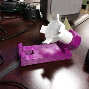 May include: A purple 3D printed phone stand with a white plastic phone holder attached to the side. The stand is designed to hold a phone in a vertical position.