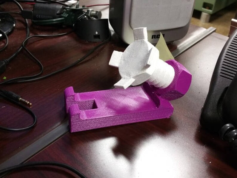 May include: A purple 3D printed phone stand with a white plastic phone holder attached to the side. The stand is designed to hold a phone in a vertical position.