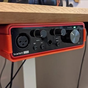 May include: A red and black Scarlett Solo audio interface mounted under a wooden desk. The interface has several knobs, buttons, and input jacks. The text "Scarlett Solo" is printed on the front.