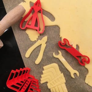 May include: A collection of red and yellow cookie cutters in the shape of tools, including pliers, wrenches, and Allen wrenches. The cutters are arranged on a sheet of dough, ready for baking.