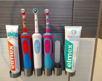 Puede incluir: Un soporte de cepillo de dientes de pared blanco para baño con cuatro ranuras para cepillos de dientes y dos ranuras para pasta de dientes. El soporte tiene cuatro cepillos de dientes eléctricos, dos azules, uno blanco y uno rosa. Hay dos tubos de pasta de dientes en el soporte, ambos son blancos con texto azul y verde que dice "elmex". Un tubo dice "sensitive" y el otro dice "junior 6-12 años".
