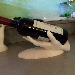 May include: White ceramic hand holding a bottle of red wine. The bottle label reads "KEG 0.75 L".