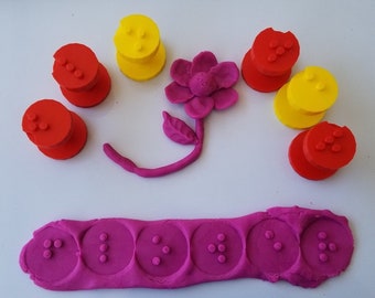 May include: A pink play dough flower with a stem and leaves, surrounded by red and yellow play dough circles with dots. A long strip of pink play dough with five circles with dots is at the bottom of the image.