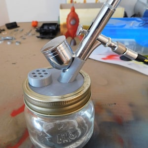 Peut inclure: Un aérographe argenté avec une buse grise et un pot en verre transparent avec un couvercle doré. L'aérographe est fixé au pot à l'aide d'un adaptateur en plastique gris. Le pot est posé sur une surface grise.