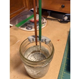 May include: A gray plastic paintbrush holder with three slots, holding two paintbrushes in a glass jar filled with water. The jar has a mesh screen at the bottom.
