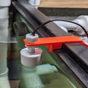 Könnte beinhalten: Ein weiß-grauer Schwimmerschalter mit einer orangefarbenen Halterung und schwarzem Draht. Der Schwimmerschalter dient zur Überwachung des Wasserstands. Das Gerät ist an einem schwarzen Metallrahmen montiert, wahrscheinlich für ein Aquarium oder ähnliches.