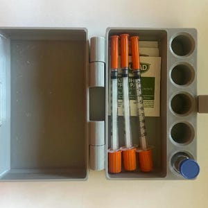 Może przedstawiać: Otwarty, szary plastikowy zestaw medyczny, zawierający strzykawki z pomarańczowymi nasadkami, chusteczki nasączone alkoholem i małą fiolkę z niebieską zakrętką. Zestaw ma pokrywę na zawiasach i przegrodę z okrągłymi otworami.