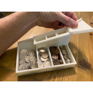 May include: A white plastic coin sorter tray featuring four distinct rectangular compartments for organizing various denominations of currency. The tray has an angled design with a hinged lid partially open, revealing stacks of silver and copper coins.