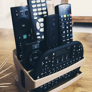 May include: A beige 3D printed remote control holder with six black remote controls inside. The holder has two levels and the remotes are arranged vertically. The remotes have buttons with various symbols and text, including "OK", "CLEAN", "LIVE", and "Rii".