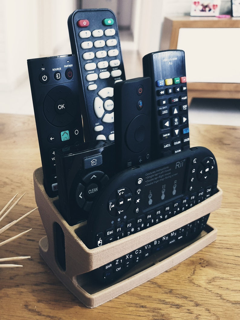 May include: A beige 3D printed remote control holder with six black remote controls inside. The holder has two levels and the remotes are arranged vertically. The remotes have buttons with various symbols and text, including "OK", "CLEAN", "LIVE", and "Rii".