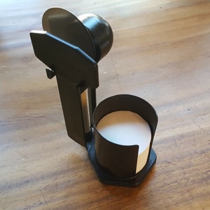 以下が含まれることがあります： 白い紙のロールが入った黒いプラスチック製のディスペンサー。ディスペンサーは丸いトップと側面にハンドルが付いています。