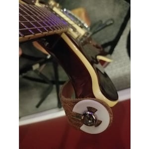 Puede incluir: Primer plano de una correa de guitarra de cuero marrón con un botón de correa de plástico blanco. La correa está unida a una guitarra.