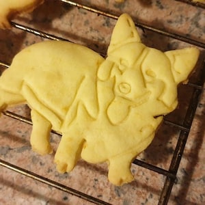 Puede incluir: Una galleta de azúcar amarilla con forma de perro corgi. La galleta tiene un diseño detallado de la cara y el cuerpo de un corgi.
