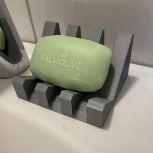 May include: A gray 3D printed soap dish with a bar of Palmolive soap on top. The soap dish has a series of ridges on the bottom to help drain water.
