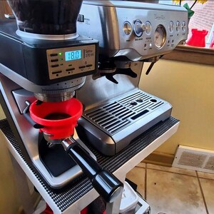 May include: A stainless steel espresso machine with a black grinder and a red portafilter. The machine has a digital display showing a temperature of 188 degrees. The image also shows a window with red accessories.