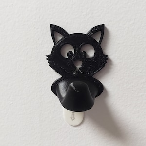 Puede incluir: Gancho de pared negro con forma de gato con una base de plástico blanco. El gancho está diseñado para parecer un gato de dibujos animados con ojos grandes y una sonrisa.