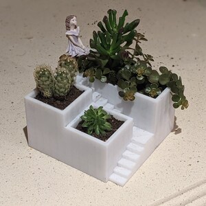 May include: White ceramic planter with three tiers, each containing a different type of succulent plant. A small porcelain figurine of a girl is placed on the top tier.
