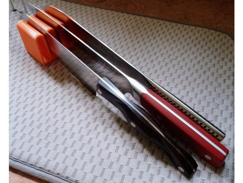 May include: Three kitchen knives are stored in a vertical orange magnetic knife holder. The knives have black, red, and silver handles.