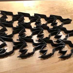 Puede incluir: Un patrón geométrico impreso en 3D de color negro hecho de formas que se entrelazan. El patrón está formado por unidades repetitivas de una forma curva que se asemeja a un ala de pájaro.