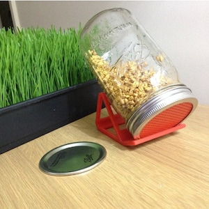 Op de afbeelding: Een rode plastic standaard houdt een glazen pot gevuld met ontkiemende granen vast. De pot is schuin geplaatst en het deksel ligt er bovenop. Een apart deksel ligt op de tafel naast de standaard.