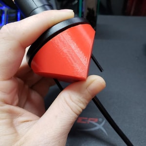 May include: A red and black 3D-printed object is held in a hand. The object has a curved, angular shape with a bright red surface and a black rim. A black cable extends from the object.