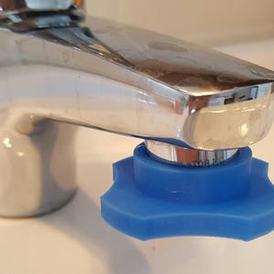May include: A close-up of a chrome faucet with a blue, star-shaped handle. The handle is attached to the base of the faucet. The faucet is on a white surface.