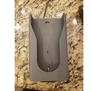 May include: A gray plastic wall mount for a phone or other device. The mount has a slot for a charging cable and a hole for a headphone jack.