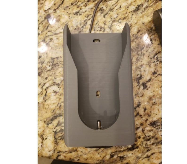 May include: A gray plastic wall mount for a phone or other device. The mount has a slot for a charging cable and a hole for a headphone jack.