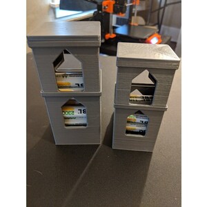 May include: Two gray 3D printed towers with a square base and a house-shaped top. Each tower has three levels and a window opening on each level. The windows are filled with rolled-up bills of money.