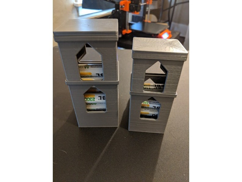 May include: Two gray 3D printed towers with a square base and a house-shaped top. Each tower has three levels and a window opening on each level. The windows are filled with rolled-up bills of money.