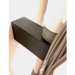 Peut inclure: Un support en plastique noir pour une baguette d'aspirateur, avec une pièce en caoutchouc gris sur le dessus. Le support est fixé à un poteau blanc avec un faisceau de cordons gris.