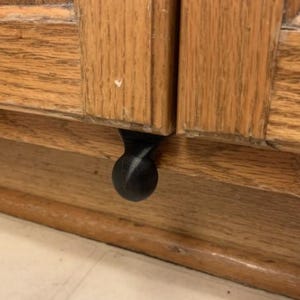 Op de afbeelding: Close-up van een houten kast met een zwarte, ronde knop. De kastdeuren zijn gemaakt van lichtbruin hout met zichtbare nerf. De knop is gecentreerd op de onderste rand van de kast.