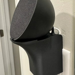 Puede incluir: Un altavoz negro montado en una toma de corriente de pared con una tapa de toma de corriente eléctrica blanca. El altavoz está inclinado hacia arriba y tiene una rejilla de tela.